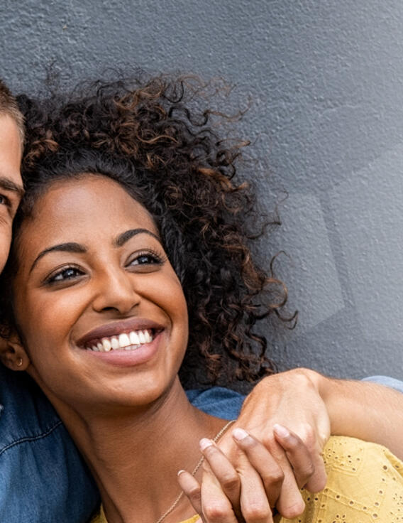 Un homme serre dans ses bras une femme, tous deux souriants, le dos contre un mur.