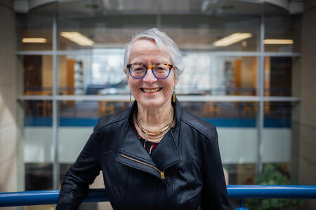 Camille, an elderly white woman with gray hair and glasses smiling. She is inside a building leaning on a railing with interior windows far behind.