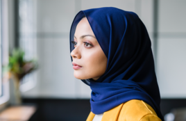 Femme portant un hijab bleu debout près d'une fenêtre donnant sur l'extérieur.